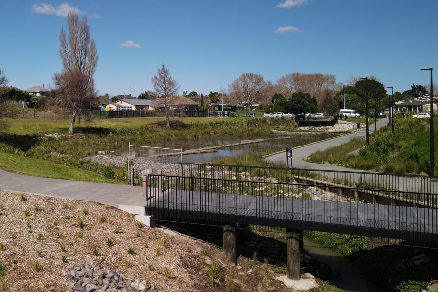 Sunny park scene with a pedestrian bridge over a stream, bare trees, and houses in the background, likely in Gisborne.