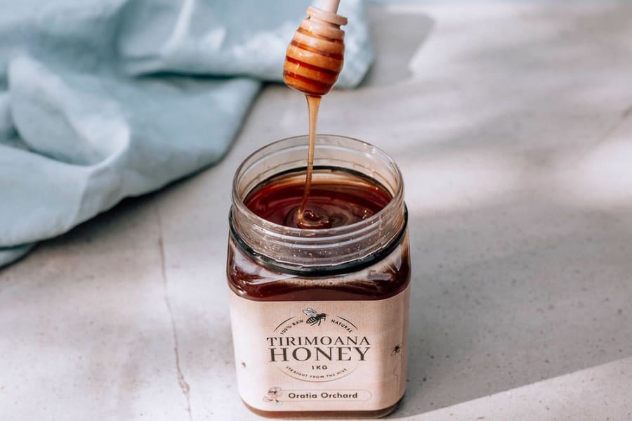 Honey dipper dripping raw Tirimoana honey into a square glass jar, with a light blue cloth on a countertop. Artisan honey from Oratia Orchard.