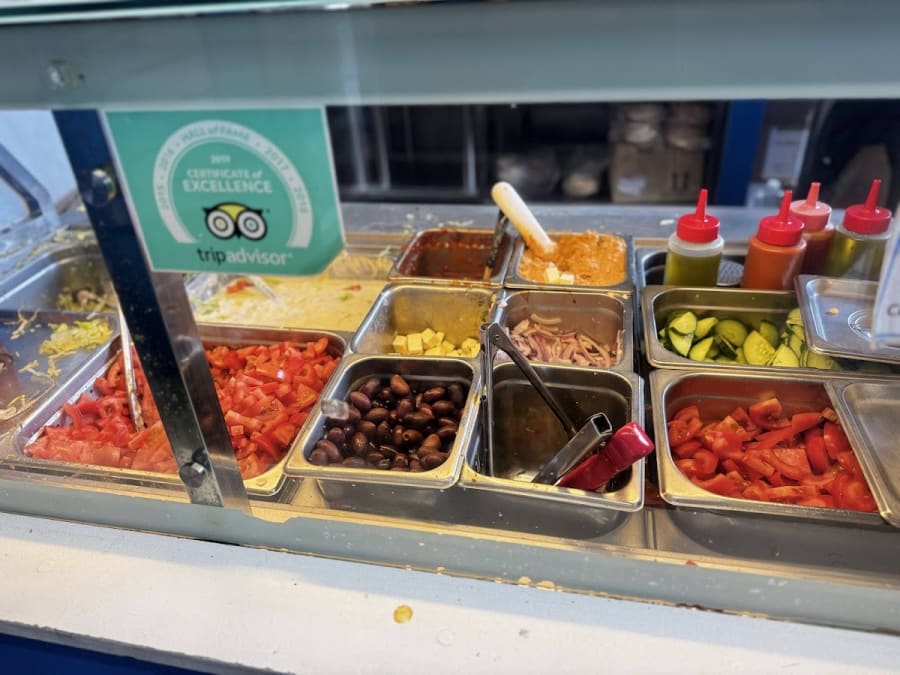 Fresh Greek food ingredients like diced tomatoes, olives, and cucumbers in a counter display, with a Tripadvisor Certificate of Excellence visible.