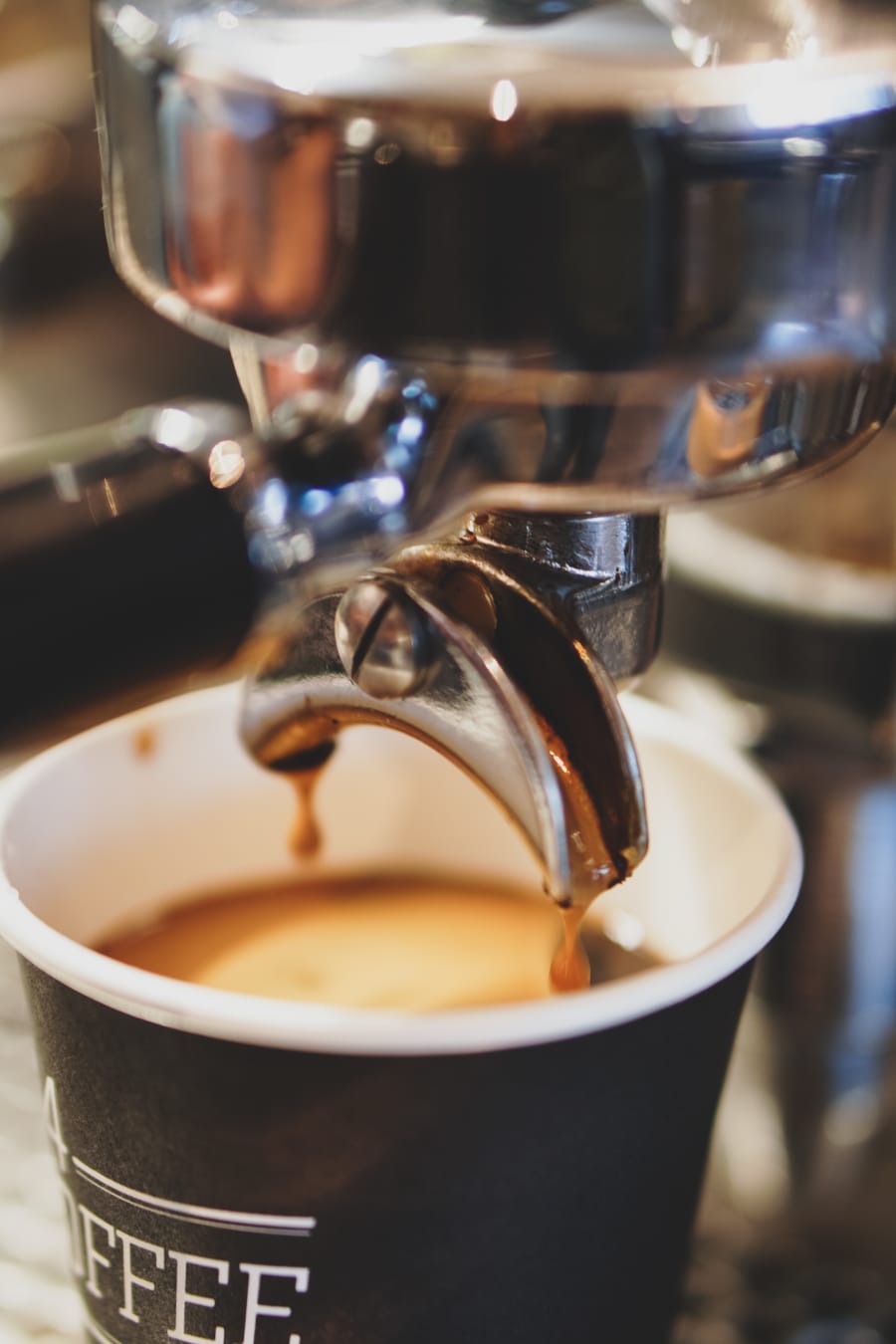 Close-up of an espresso machine portafilter dripping rich coffee with golden crema into a black takeaway cup.