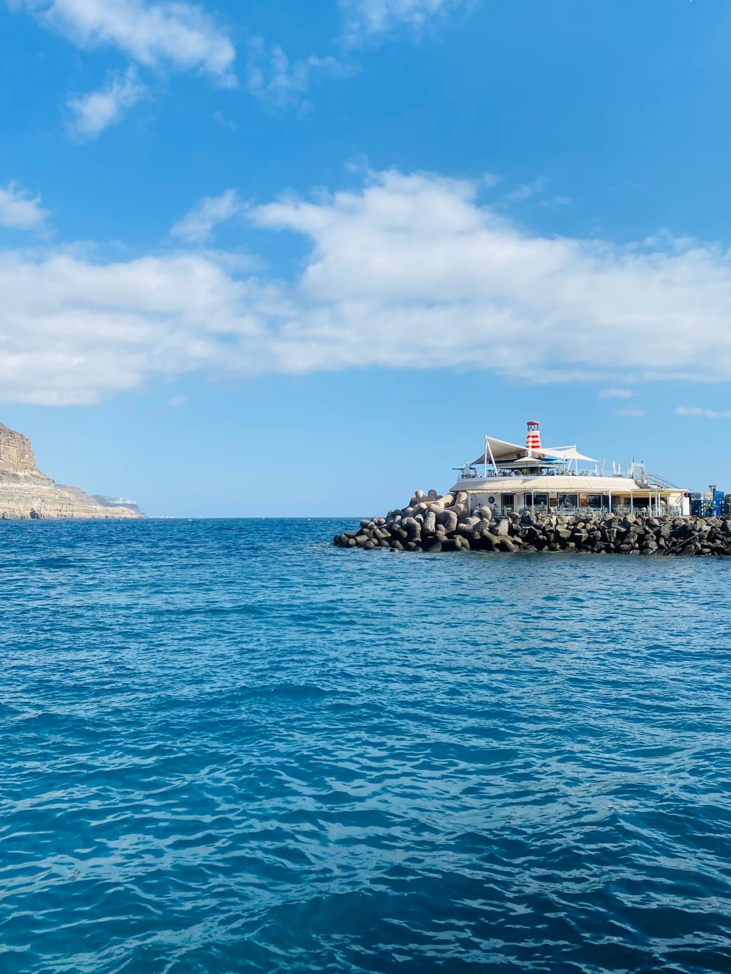 The harbour wall at Puerto de Mogán, with a small striped lighthouse at its end and cliffs along the coast