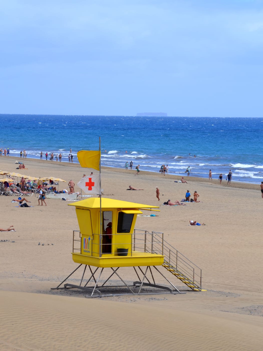 A yellow lifeguard post on the long beach at Maspalomas, with people walking along the surf