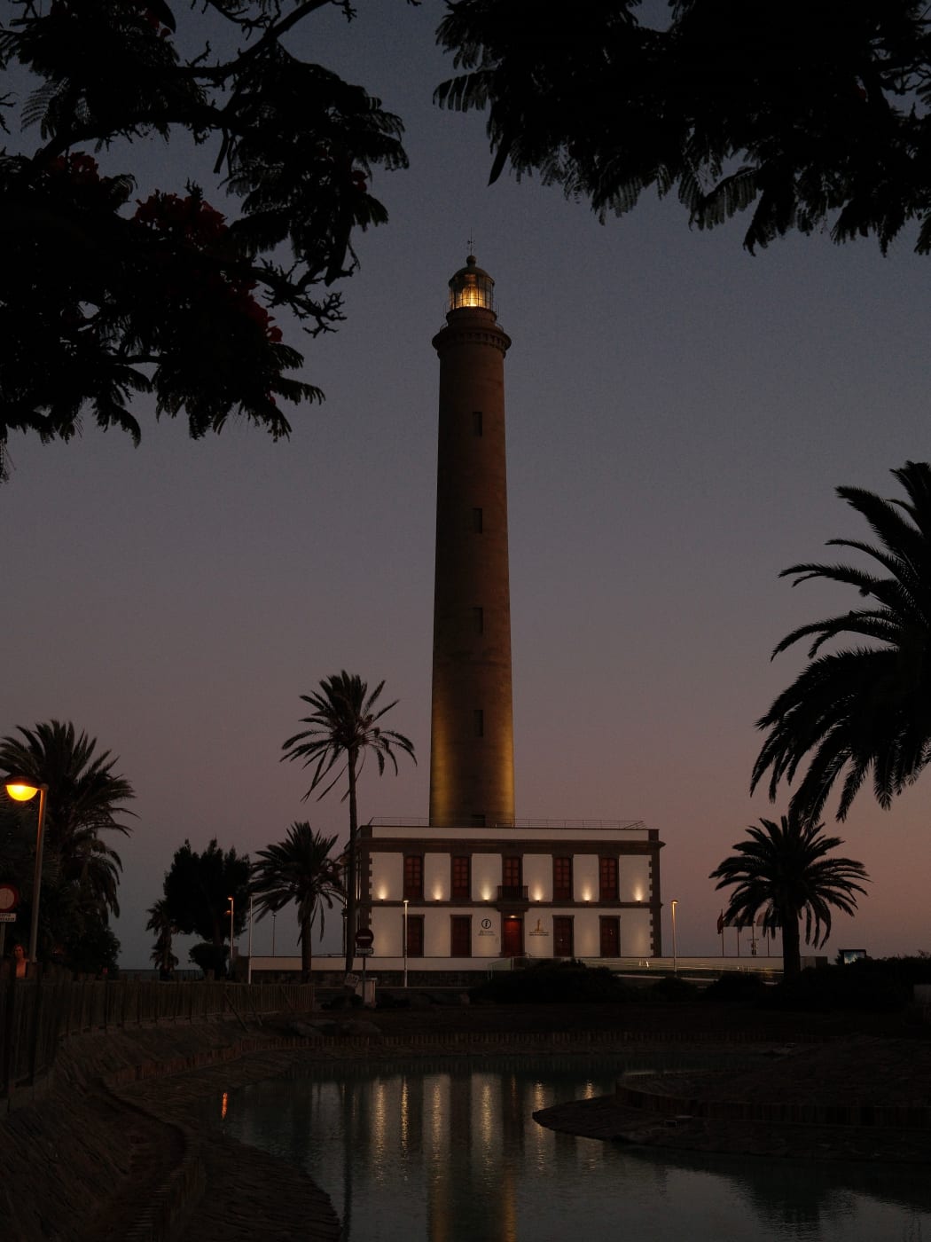The Faro de Maspalomas lit up at dusk behind palm trees and a pool of water