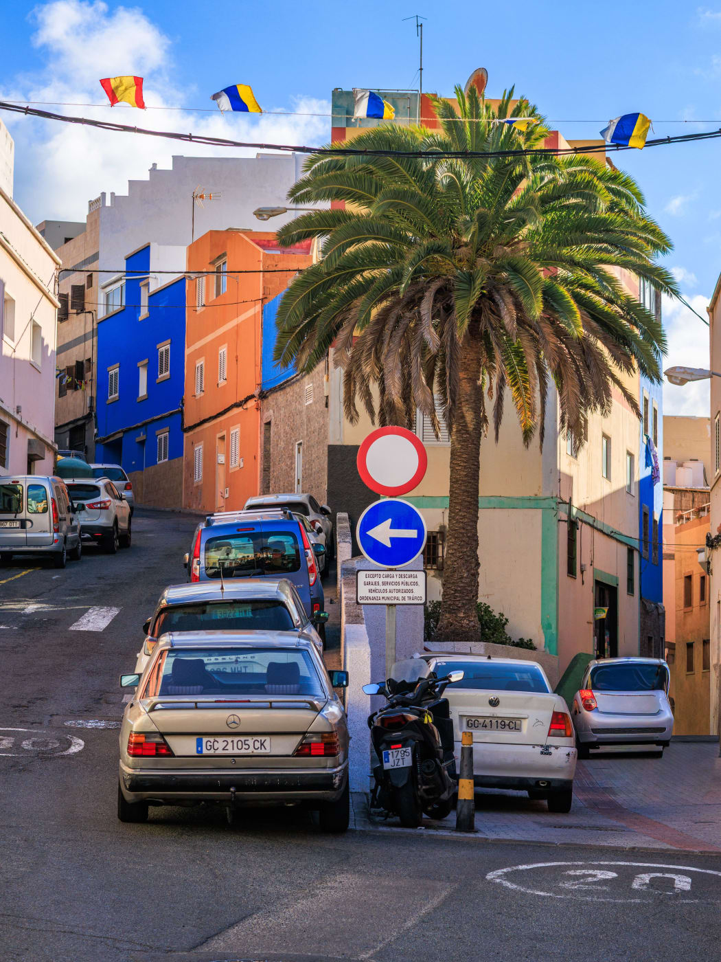 Cars parked along a steep street in Las Palmas under a palm, with a 20 speed limit painted on the road