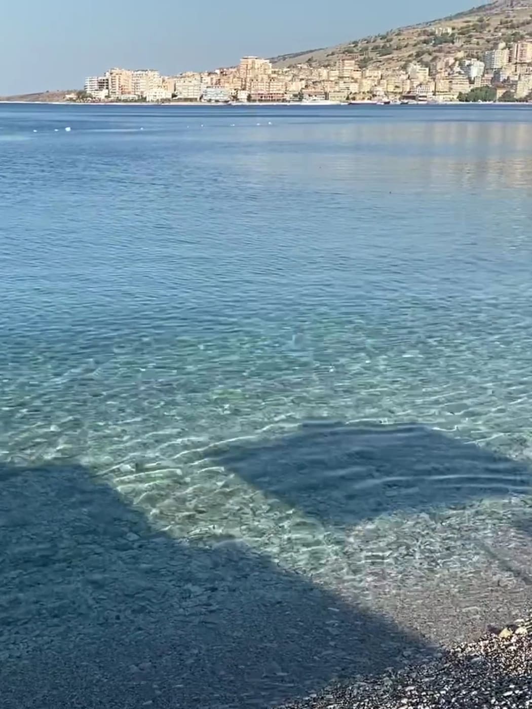 Eau claire et peu profonde sur les galets du rivage du Buzë Hotel, sur la route côtière au sud, avec la ville de Saranda de l'autre côté de l'eau