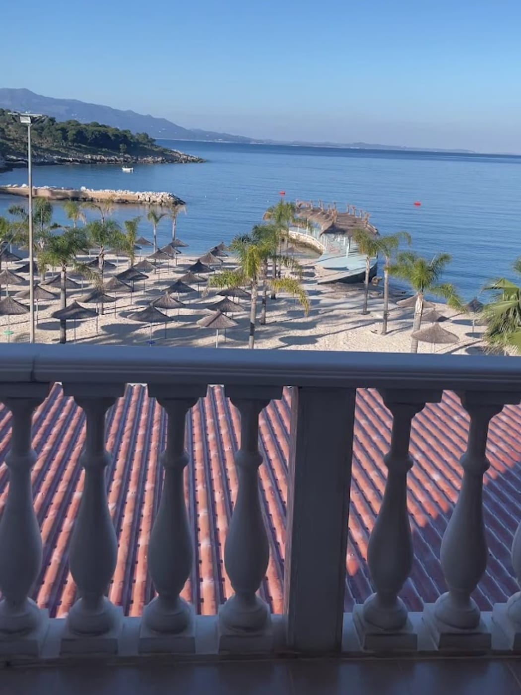 Parasols en paille et palmiers sur la plage d'hôtel à l'ouest de la promenade, vus depuis le balcon d'un hôtel Mucobega au-dessus d'un toit de tuiles