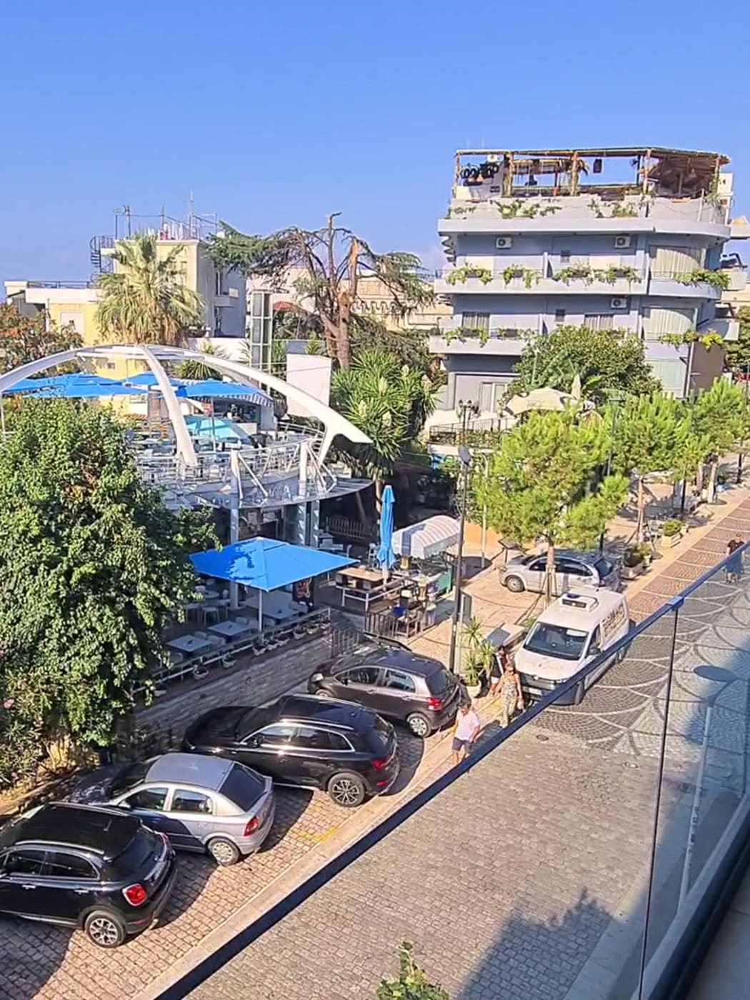 Palmiers et voitures garées dans une rue du centre de Saranda, vus depuis le balcon d'un hôtel, avec la baie au-delà