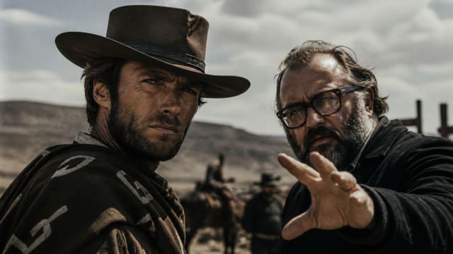 Clint Eastwood in cowboy attire stands beside director Sergio Leone in a dusty western landscape, both staring intensely into the distance beneath a dramatic cloudy sky.