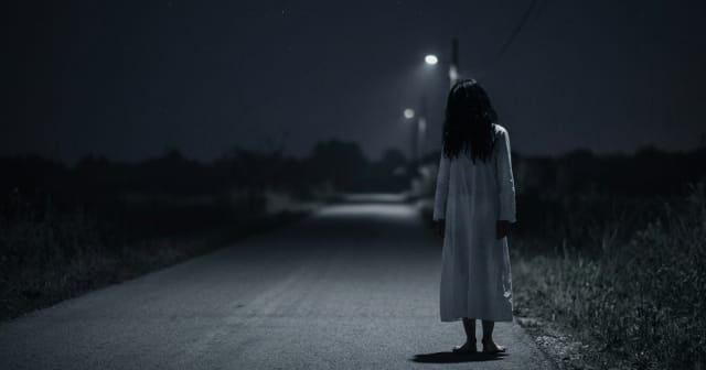 A woman in a white dress stands alone on a deserted road at night, viewed from behind under dim streetlights, with her feet unnaturally turned backward.