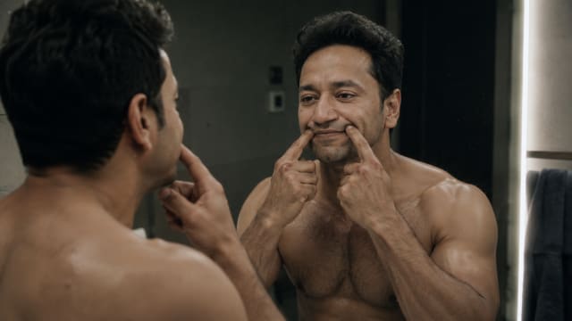 Shirtless muscular man forcing a smile in a dark bathroom mirror at night, his exhausted eyes revealing sadness and emotional strain beneath performative composure.