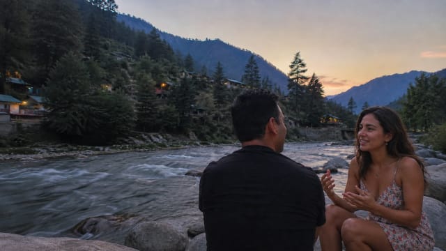 A man and his Andalusian friend sit beside the Parvati River in Kasol at dusk, discussing literature, history, language, and modern internet culture against a backdrop of mountains and pine forests.