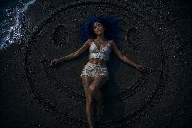 A young woman with vivid blue hair lies on wet beach sand at night inside a giant hand-drawn smiley face, photographed from directly overhead as moonlight and gentle ocean waves create a surreal, contemplative scene about happiness, loneliness, and the masks people wear.