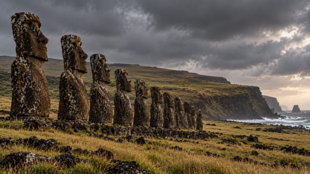 Rows of weathered stone statues facing a windswept grassy slope on a remote island under a heavy grey sky.