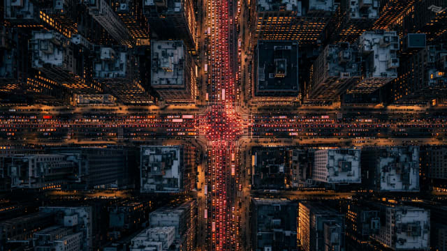 Cinematic aerial view of a congested Manhattan-style city intersection at dusk, with dense traffic, glowing red taillights, illuminated skyscrapers and geometric urban streets illustrating the Braess paradox and the mathematics of gridlock.