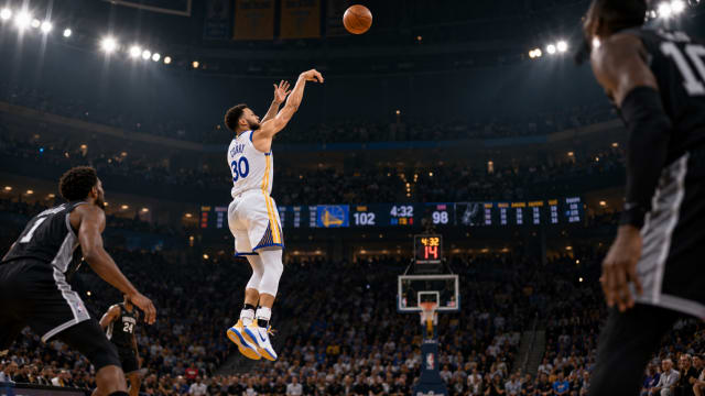 A basketball player in mid jump releases a shot beneath bright arena lights while a scoreboard of scattered numbers glows behind the crowd.