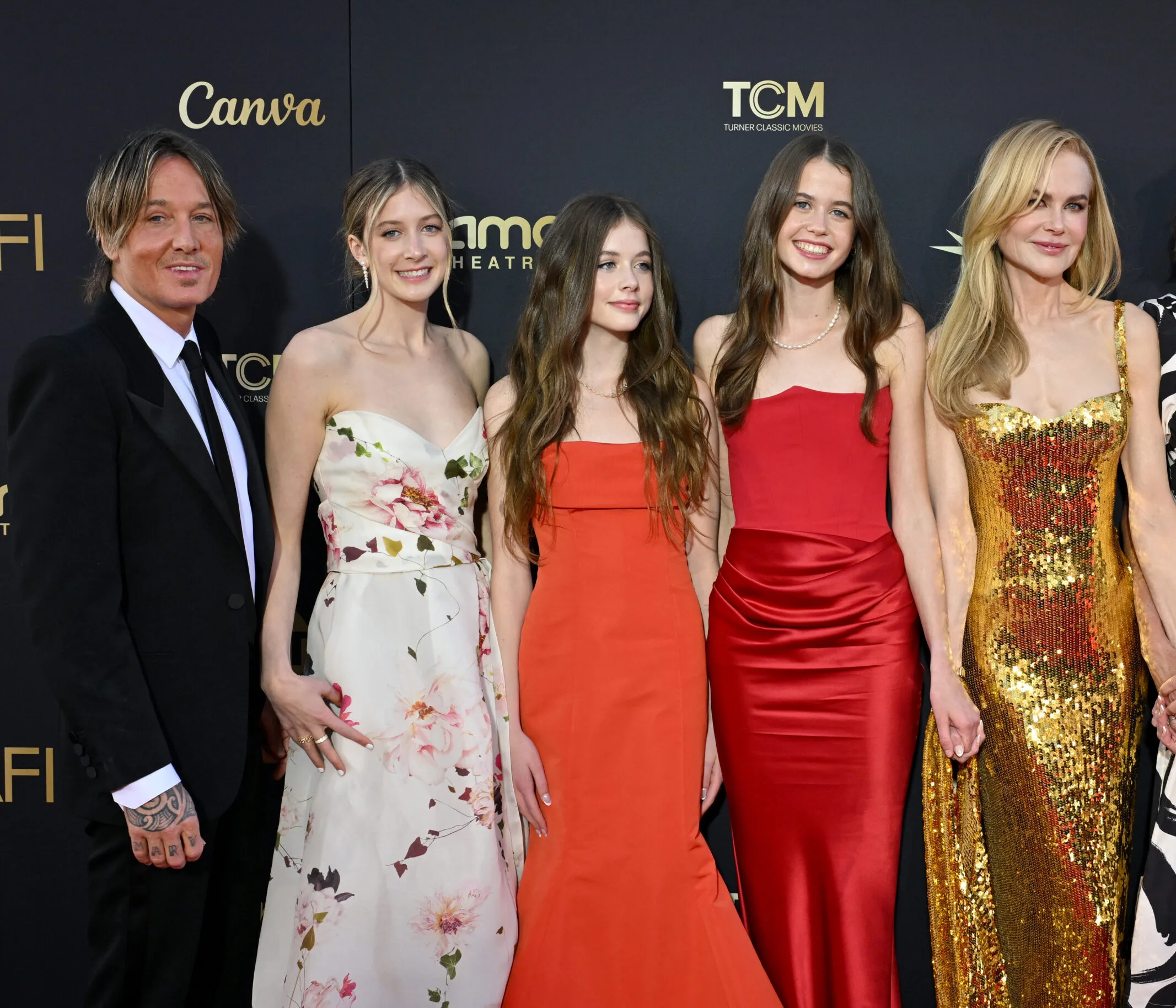 Keith Urban in a black suit, Sunday Rose in a floral gown, Faith Margaret in an orange dress, and Nicole Kidman in a gold sequined gown, posing together on a red carpet backdrop.