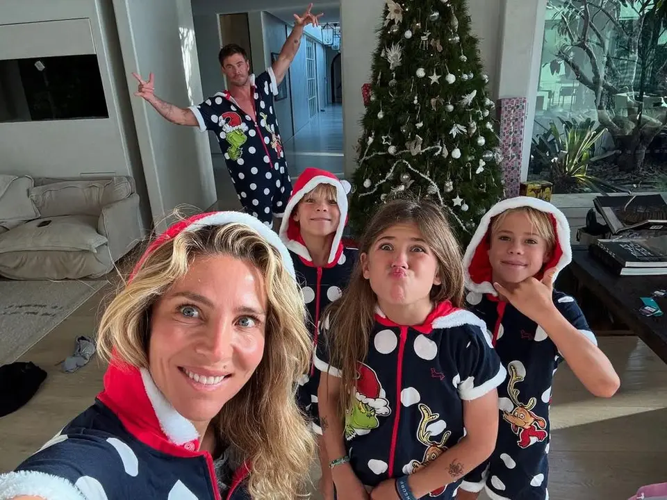 Elsa Pataki with her three kids standing in front of a Christmas tree with Chris Hemsworth in the background raising both arms making 'Rock-on' sign with hands, all are in matching pyjamas