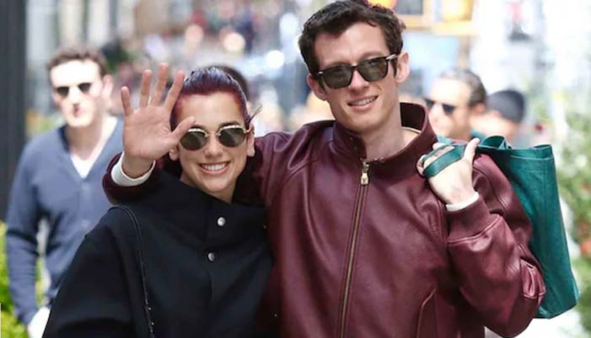Dua Lipa and Callum Turnder wearing sunglasses walk together on a sunny street, waving at the camera with crowds in the background.