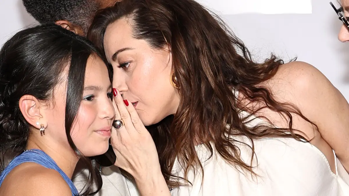 Aubrey Plaza wearing white dress whispering something to young girl in blue dress