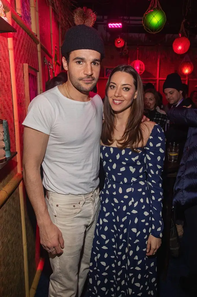 Aubrey Plaza standing with partner Christopher Abbot. Aubrey wearing a blue dress with white pattern and Christopher ios wearing white T-shirt, cream pants and a blue backwards P cap