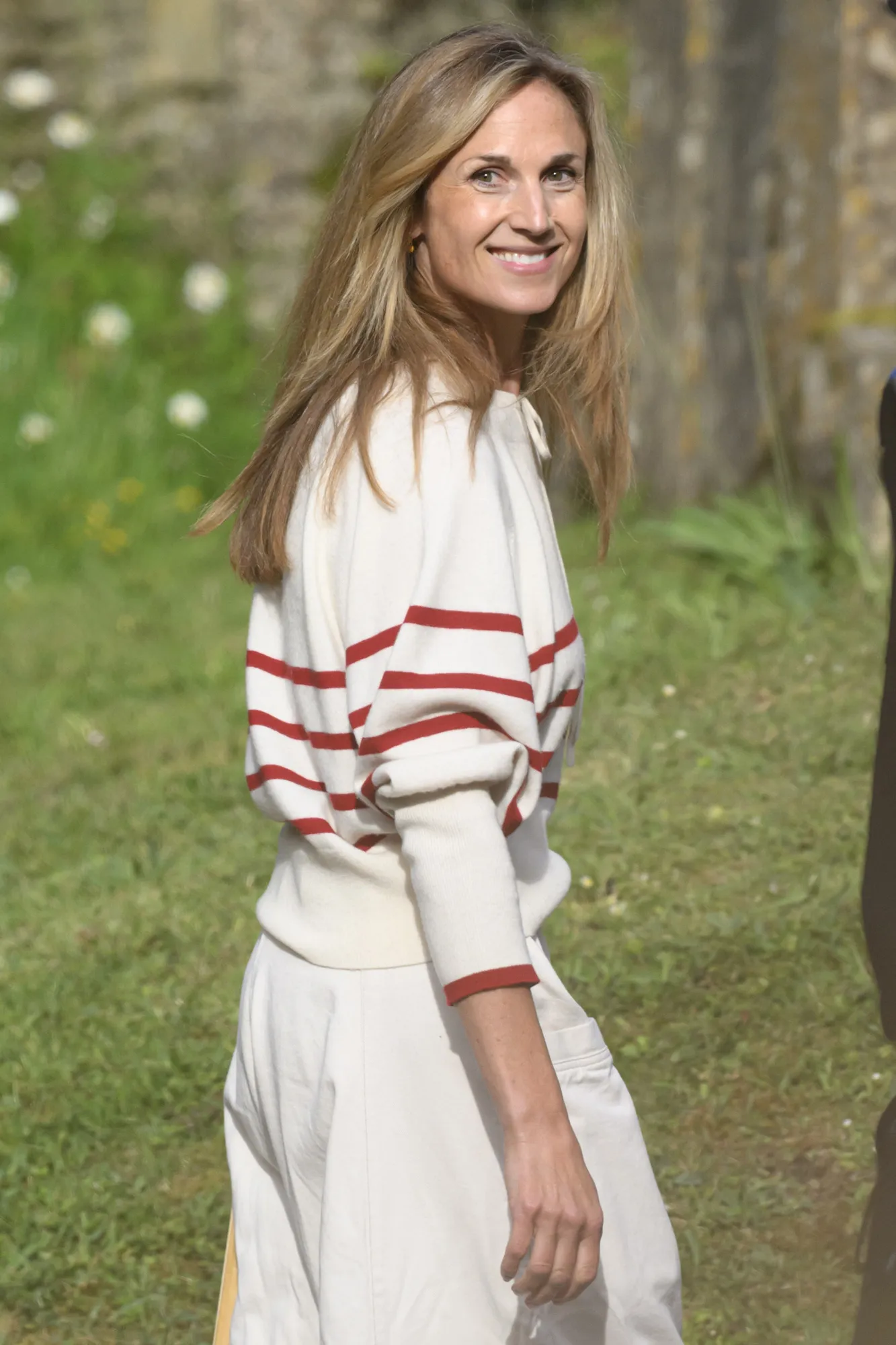 A smiling Harriet Sperling in a white and red striped sweater with white trousers, standing outdoors on a green lawn.