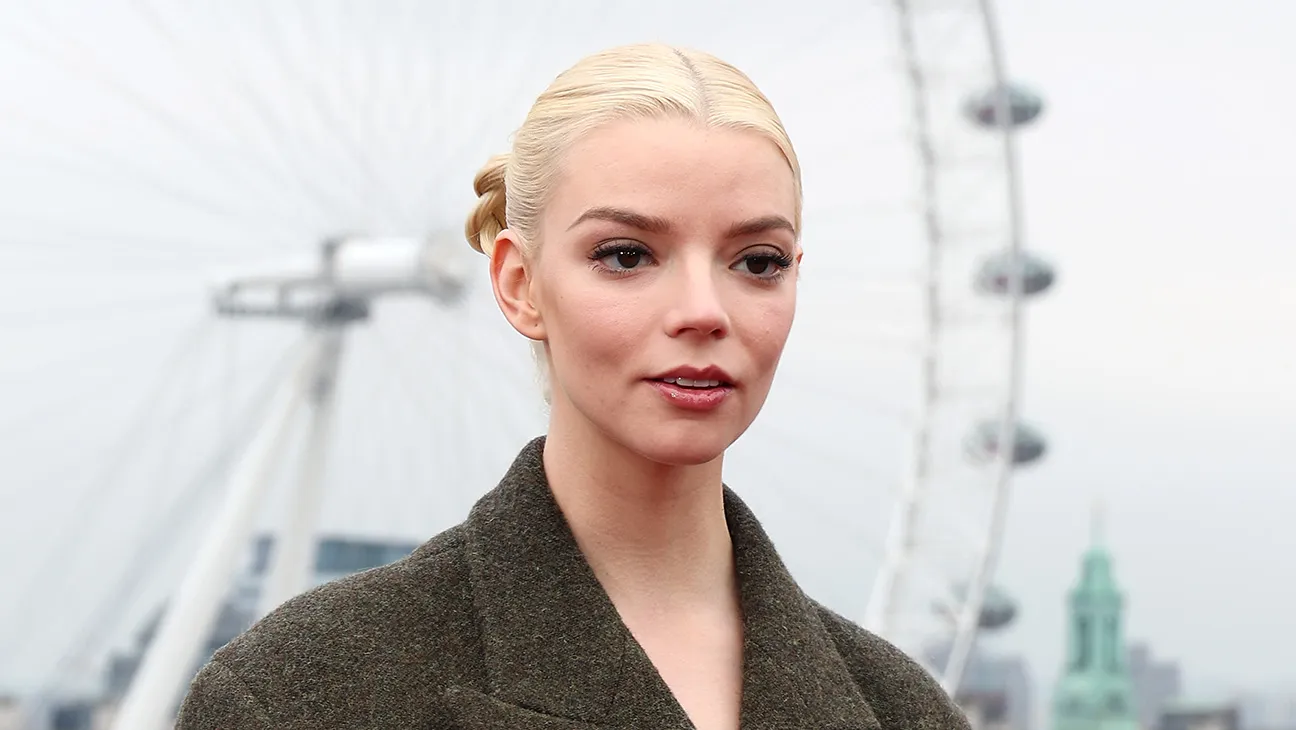 Anya Taylor Joy with slick back blonde hair wearing a green coat stands in front of what looks like the London Eye