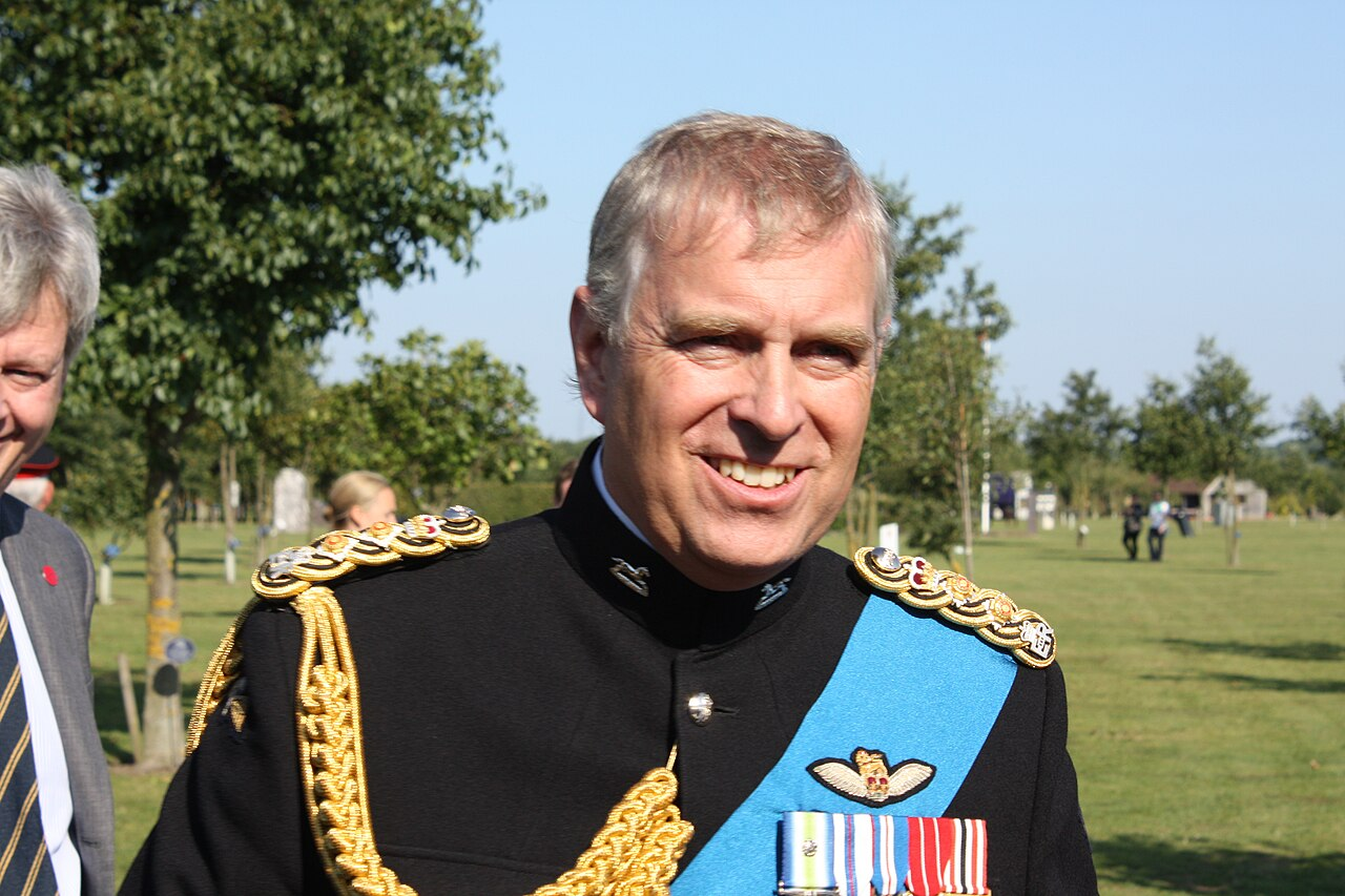 Prince Andrew in a black military dress uniform with gold epaulettes, a blue sash, and medal ribbons, standing outdoors on a sunny day.