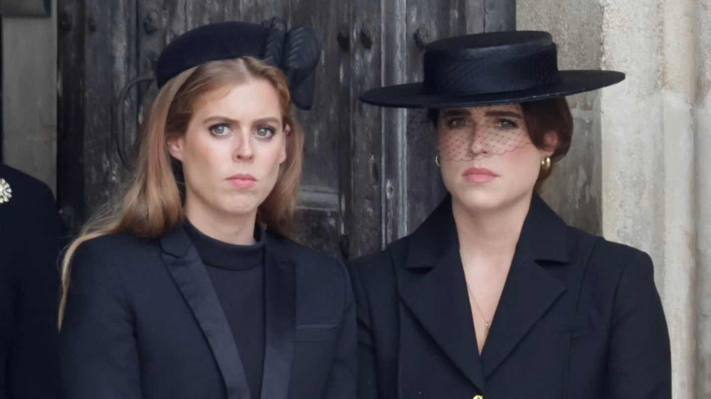 Princesses Beatrice and Eugenie dressed in black mourning attire with black hats, one wearing a veiled fascinator, standing solemnly in front of a large wooden door.