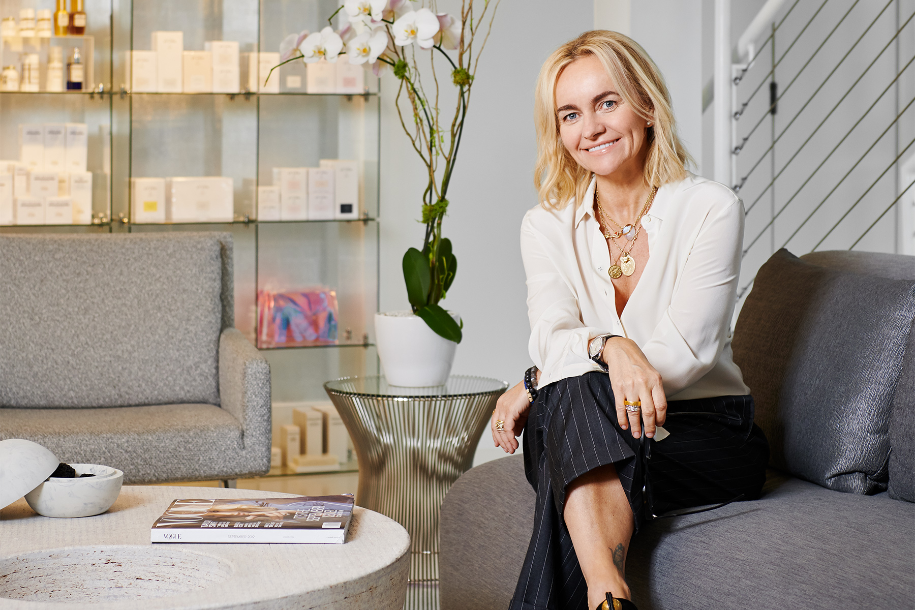 Joanna Czech in a white blouse and pinstripe trousers seated on a grey sofa in an elegant beauty salon or spa, with skincare products displayed on shelves behind her.