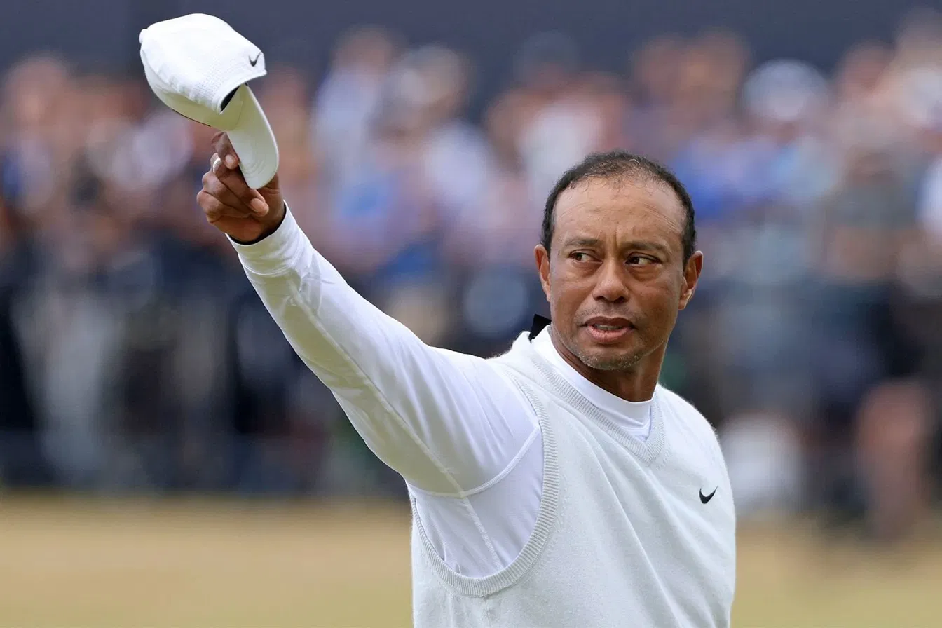 Tiger Woods in a white Nike outfit raising his cap to acknowledge the crowd at a golf tournament.