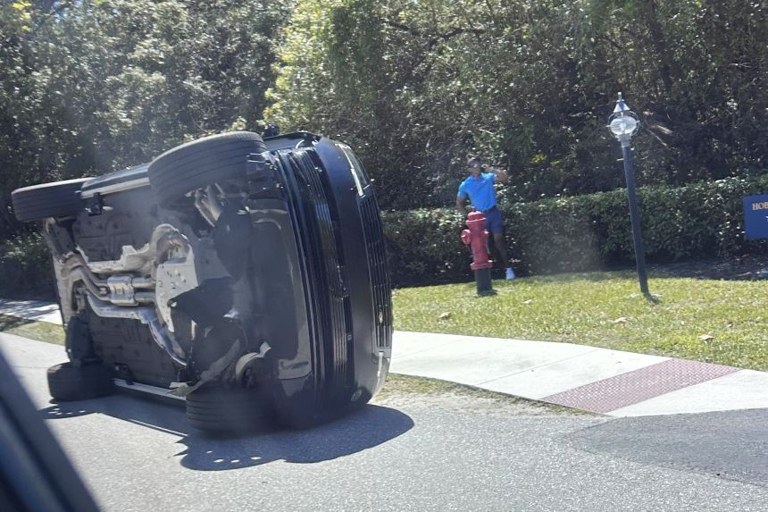 Tiger Woods' black SUV overturned on its side on a suburban street, with a bystander in a blue shirt standing nearby next to a red fire hydrant.
