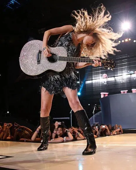 Taylor Swift performing on stage with a crystal-encrusted guitar, wearing a black sequined fringe dress and knee-high boots, hair flying dramatically under concert lights.