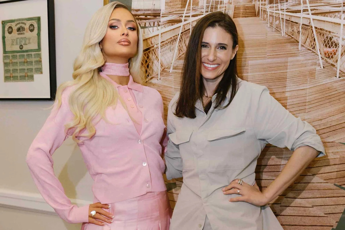 Paris Hilton and Laurie Segall pose together indoors; Paris wears a pink suit with a choker, Segall wears a beige suit, with a wooden boardwalk backdrop.