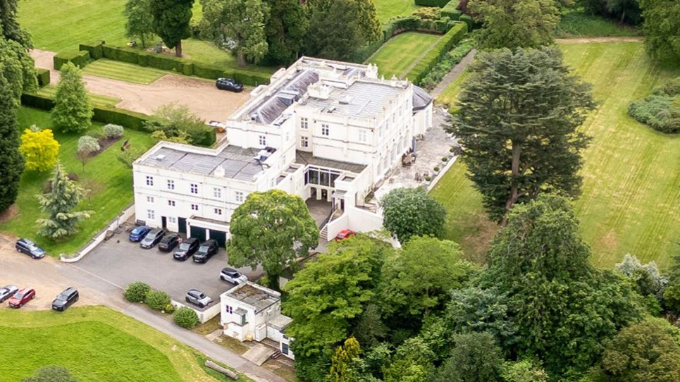 Aerial view of Royal Lodge estate in Windsor, a large white Georgian mansion surrounded by manicured lawns and mature trees.