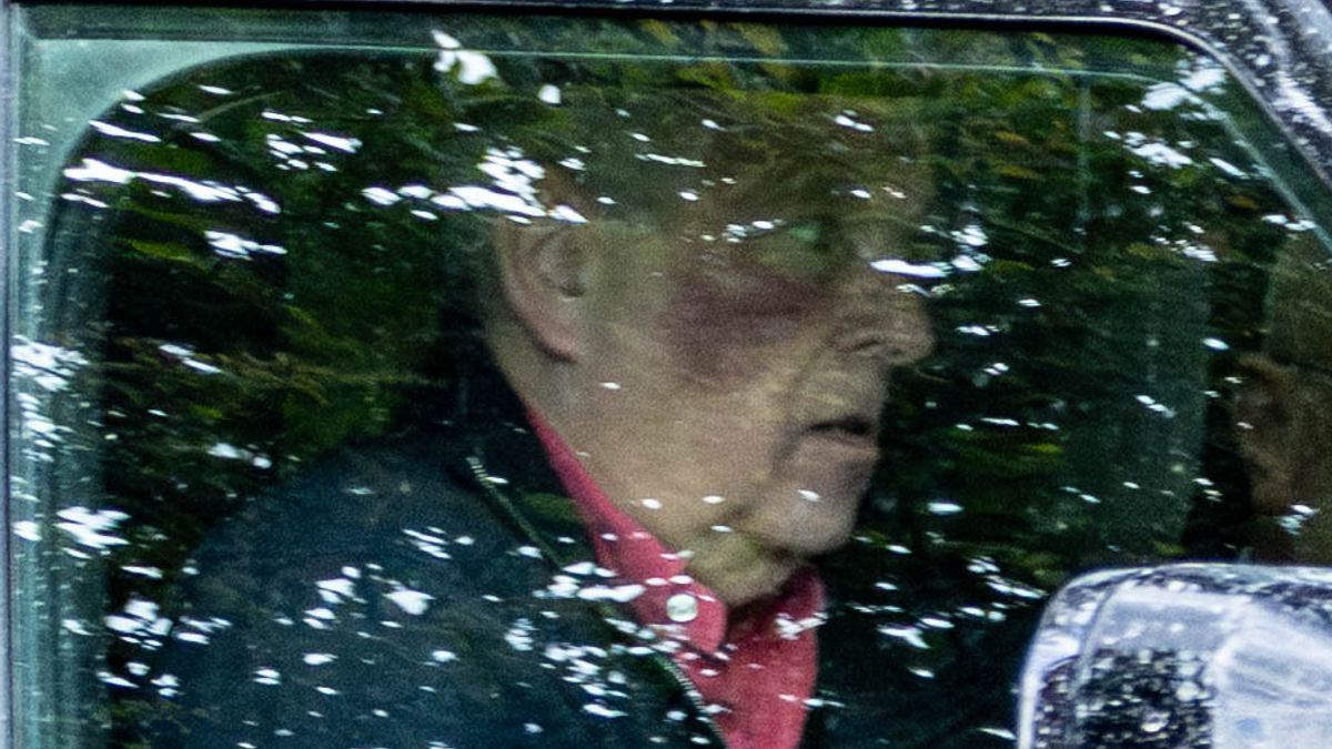 Andrew Mountbatten-Windsor. wearing a dark jacket and red garment is shown through a window with reflections of snow-covered evergreen trees.