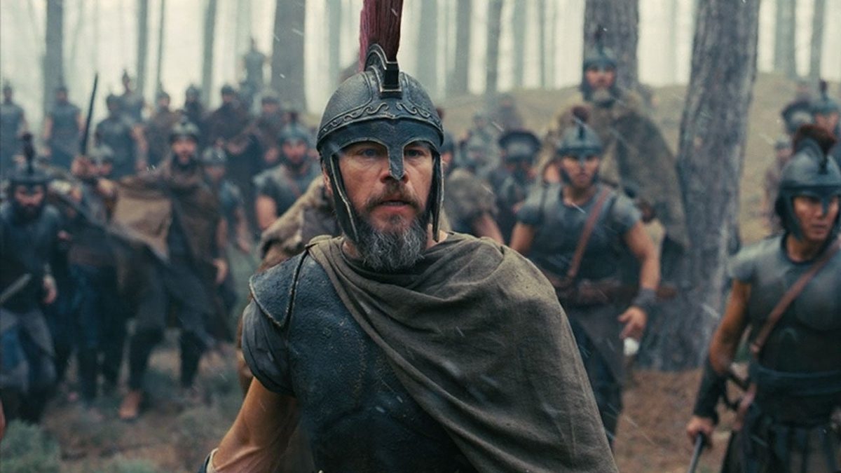 Matt Damon as Odysseus in ancient Greek warrior armour and plumed helmet, leading troops through a forest in a battle scene.