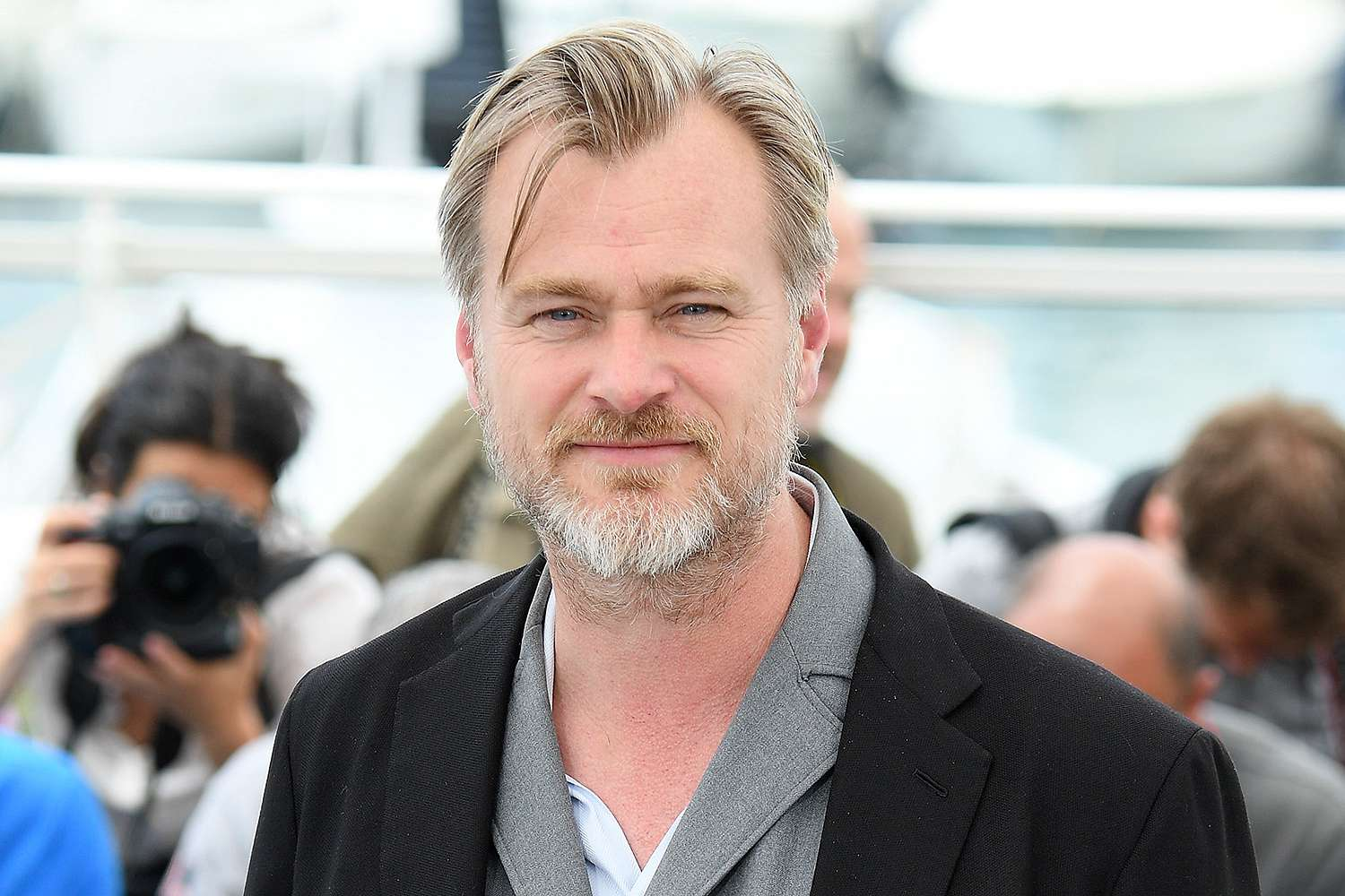 Christopher Nolan in a black blazer and grey shirt at a film event, with photographers visible in the background.
