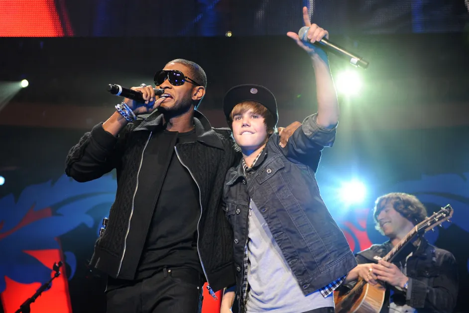 Usher and a young Justin Bieber performing together on stage, both holding microphones under concert lighting.