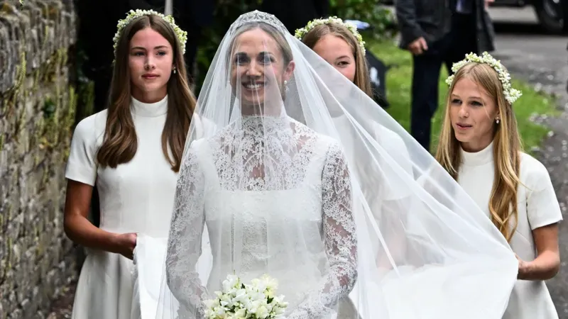 Harriet Sperling in a lace wedding gown and veil, holding a white bouquet, accompanied by bridesmaids in white dresses with floral crowns.