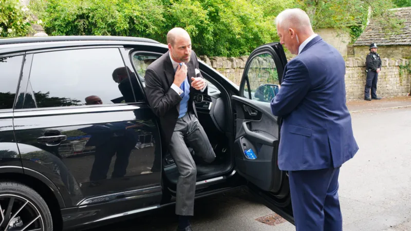 Prince William stepping out of a black SUV, adjusting his tie, greeted by an aide outside a stone-walled venue.