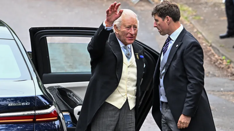 King Charles in wedding guest attire, waving as he steps out of a car at a formal event.