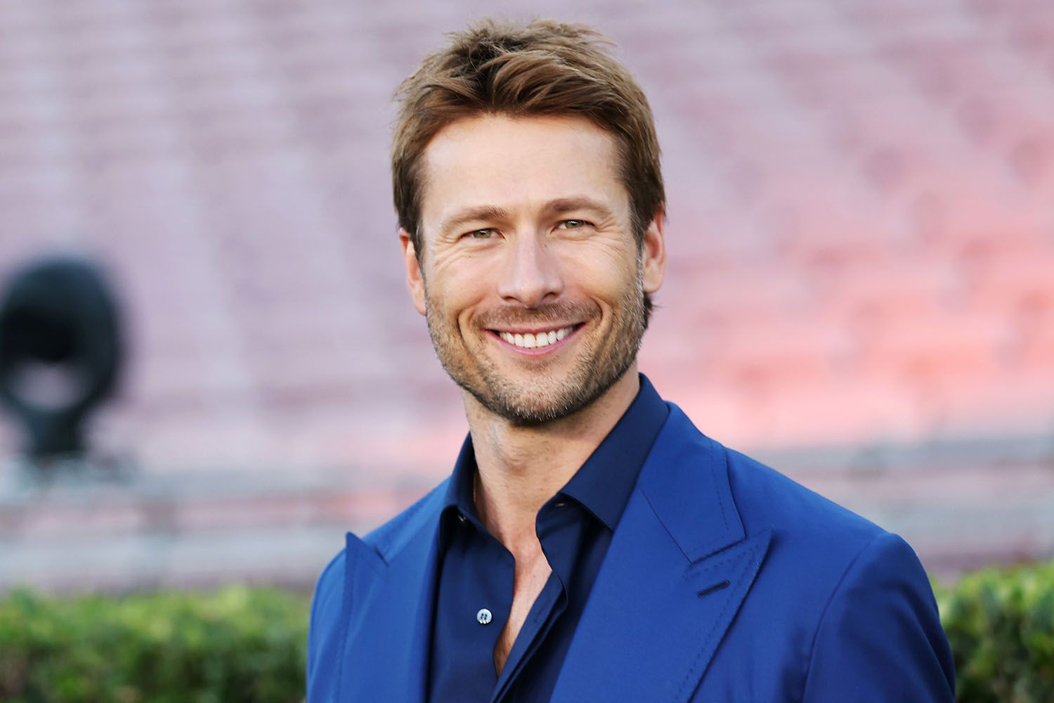 Glen Powell smiling at a public event, wearing a blue suit and matching shirt.