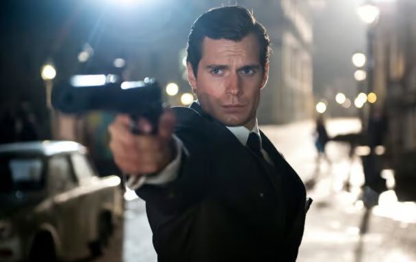 Henry Cavill in a dark suit aims a handgun directly at the camera on a nighttime city street.