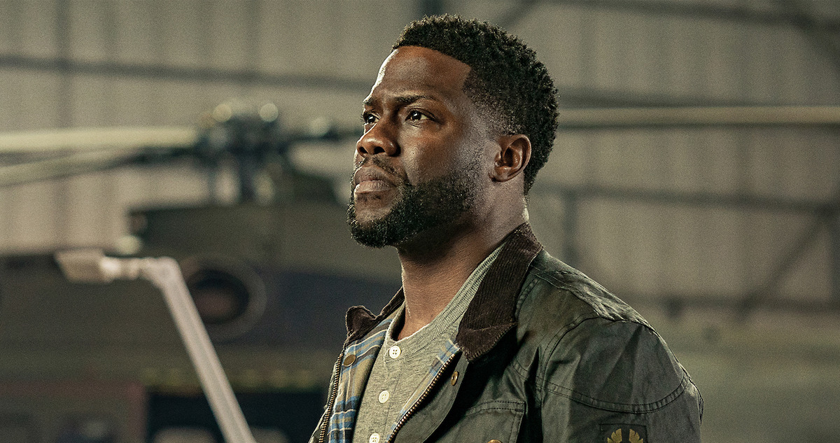 Kevin Hart in a gritty industrial setting, wearing a worn jacket, gazing upward in a dramatic film still.