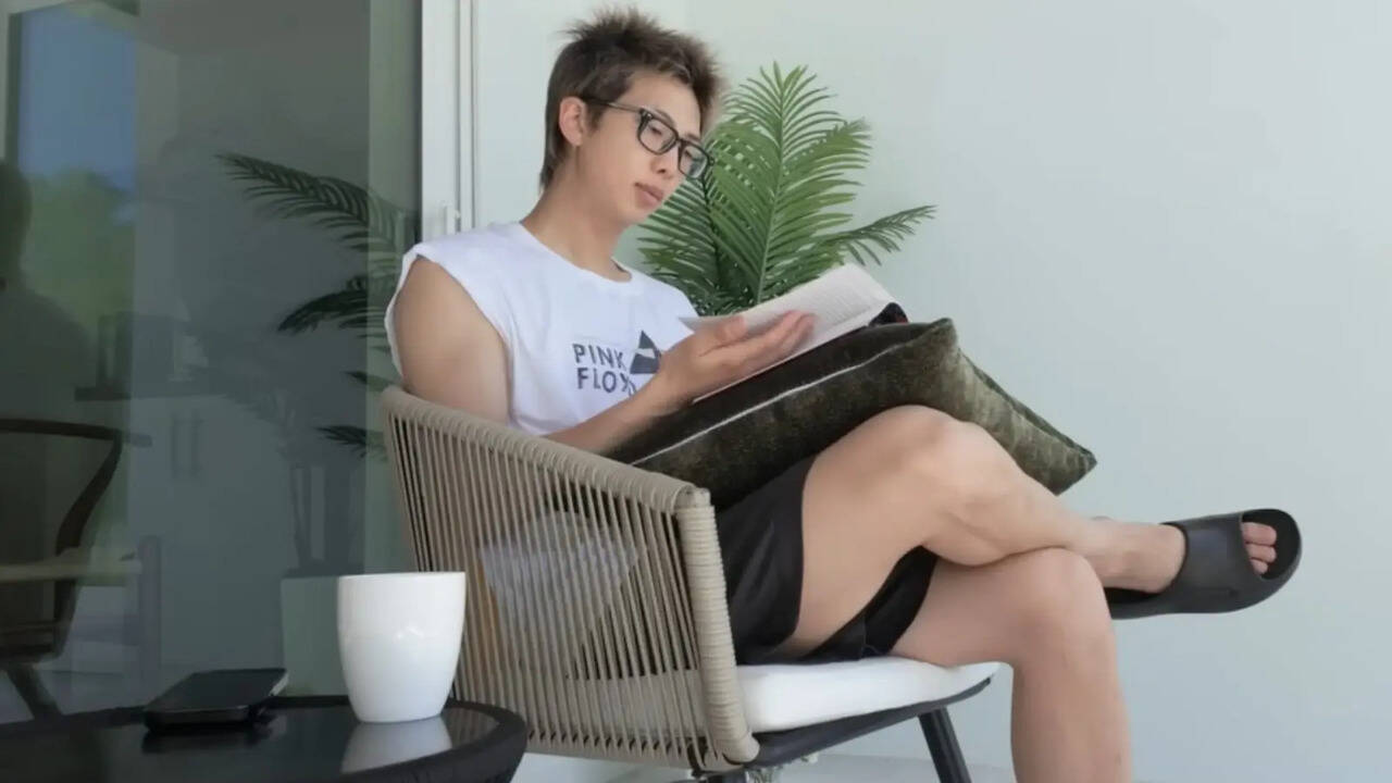 BTS band member wearing glasses and a white t-shirt sits in a modern chair reading a document, with a potted plant and coffee cup nearby.