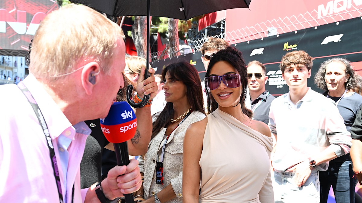 Martin Brundle tries to interview Kim Kardashian in sunglasses at a Formula 1 event, with spectators and umbrellas visible behind.