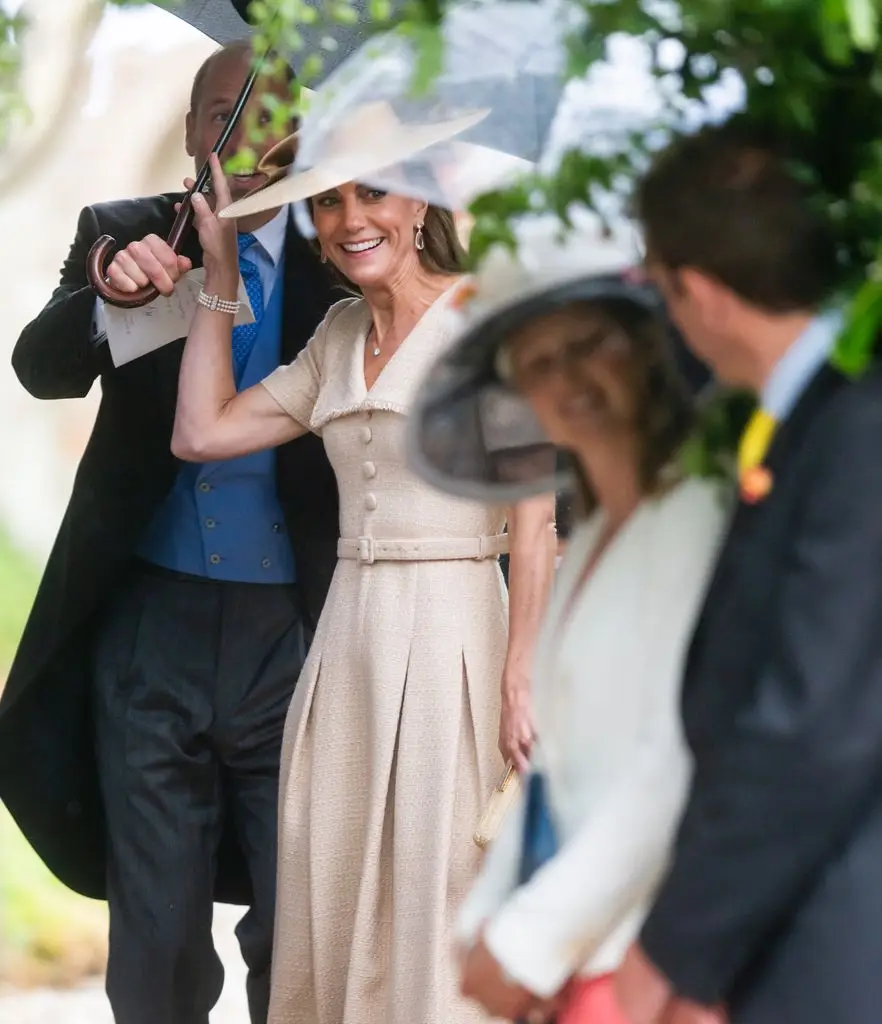 Kate Middleton in a beige belted dress and wide-brim hat, with Prince William holding a clear umbrella behind her at an outdoor event.
