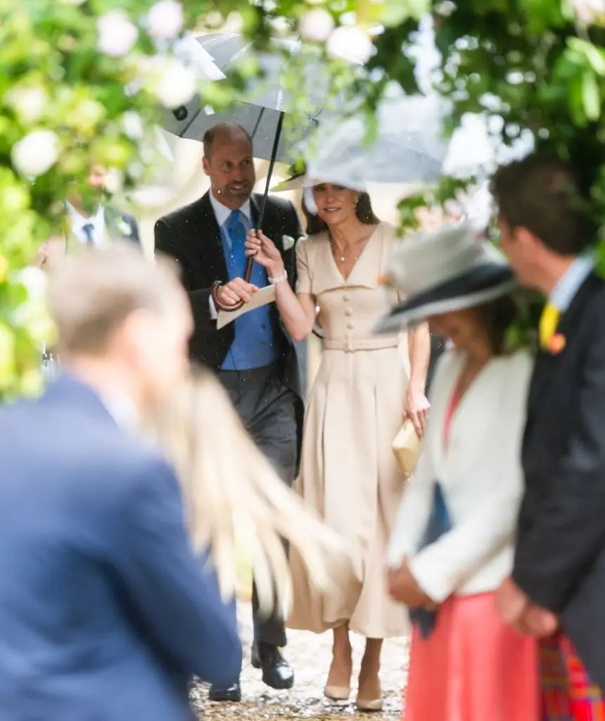 Kate Middleton in a beige belted dress and wide-brim hat, with Prince William holding sharing a clear umbrella