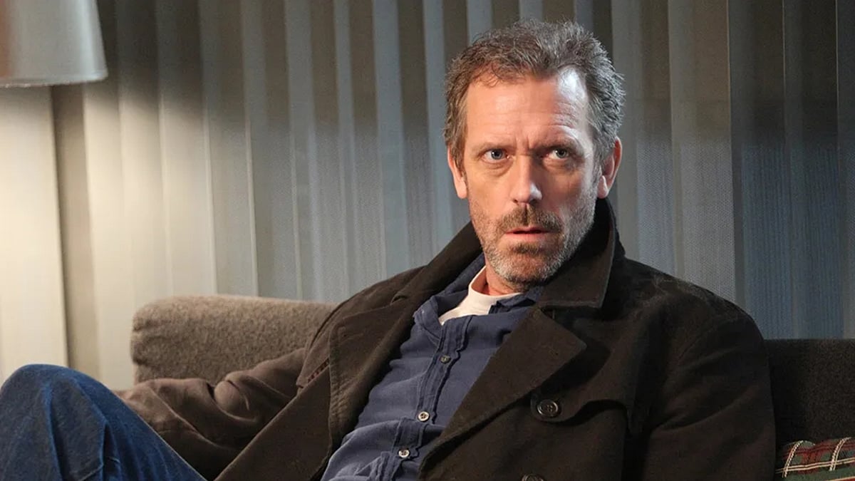 Hugh Laurie as Dr House sits on a couch wearing a dark jacket over a blue shirt, looking thoughtfully to the side, in a still from the TV Show House