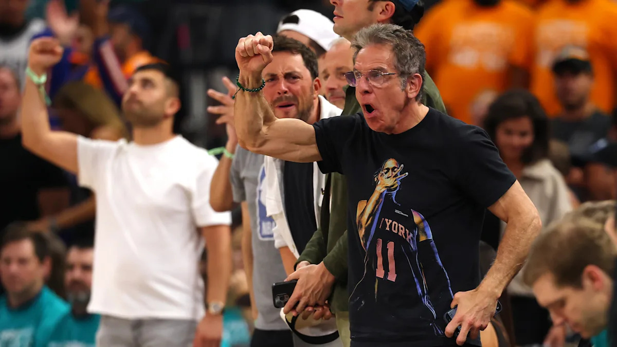 Ben Stiller in a New York basketball jersey celebrates enthusiastically at a sports game surrounded by other spectators.